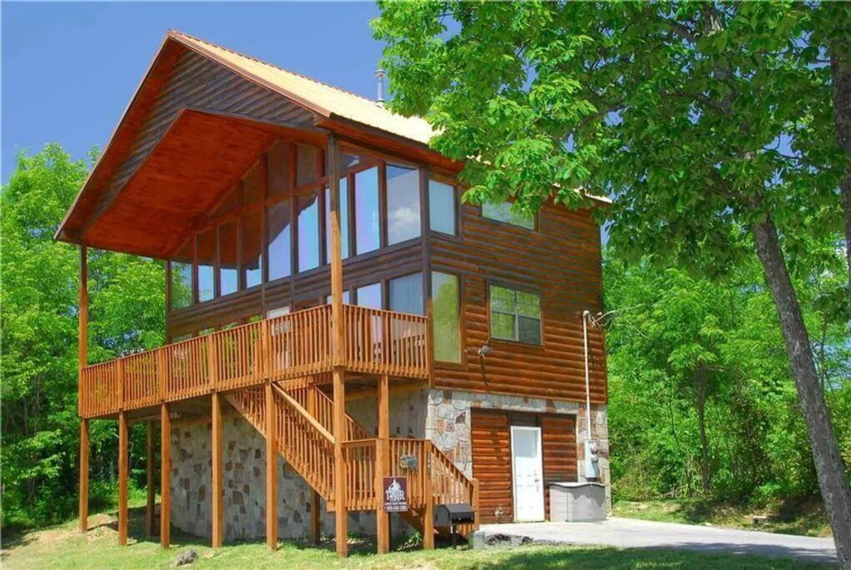 An exterior view of a wooden balcony with a stone base, surrounded by lush greenery. The balcony is equipped for gatherings, and the large windows of the cabin are prominent. The scene evokes the transition from indoor warmth to outdoor serenity, with nature all around.