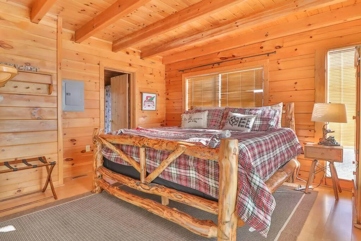 A cozy bedroom featuring a rustic log bed frame with soft plaid bedding, wooden walls, a window with blinds, and a bedside lamp. The gentle light filtering into the room creates a warm, inviting atmosphere that emphasizes comfort and privacy.