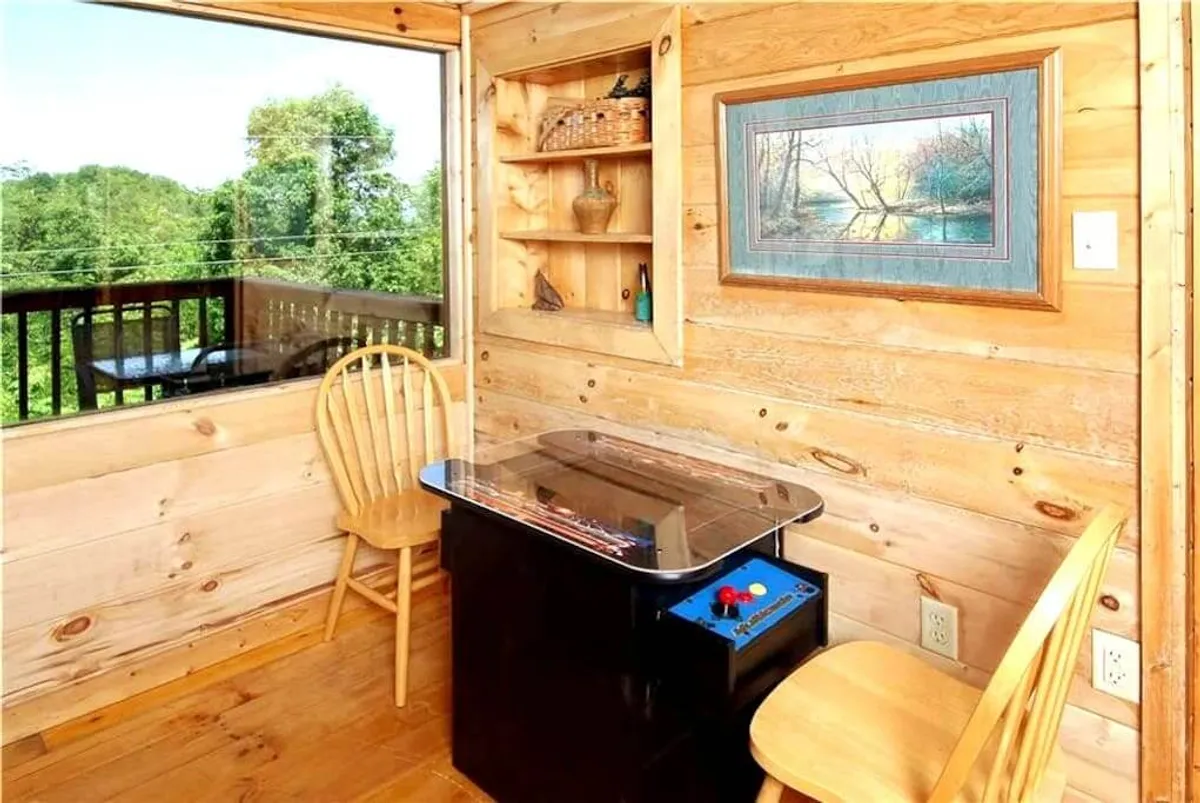 A cozy corner with wooden chairs, an arcade game table, and a large window offering views of the surrounding nature. The area is accented by framed artwork, creating a playful yet relaxed environment ideal for casual gaming and relaxation.