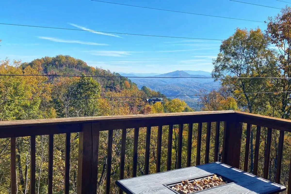 A wooden deck overlooks a stunning mountain landscape, framed by autumn foliage. A fire pit table and seating area invite guests to gather, relax, and enjoy the view—ideal for sunset moments, morning coffee, or conversations beneath the open sky.