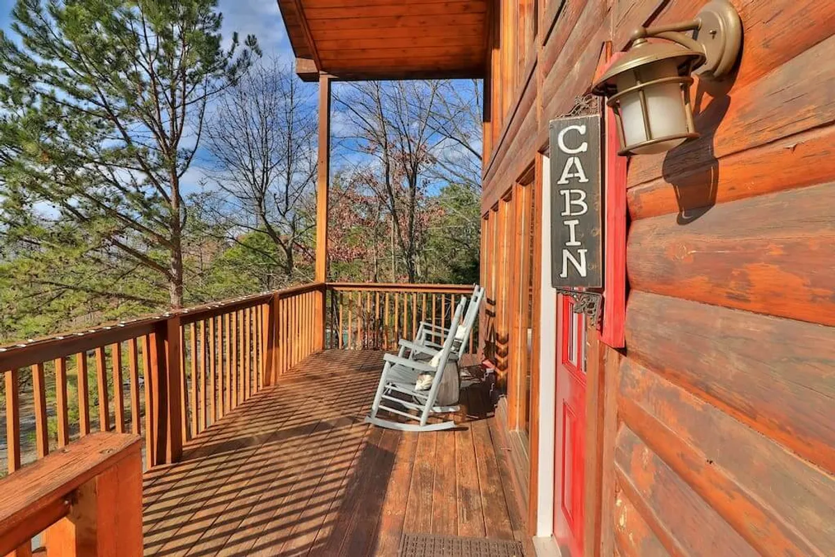 A cozy cabin porch nestled among trees, featuring a classic rocking chair, a welcoming cabin sign, and a sturdy wooden railing. The surrounding foliage and warm wood tones evoke a tranquil, private atmosphere ideal for quiet mornings or romantic evenings.