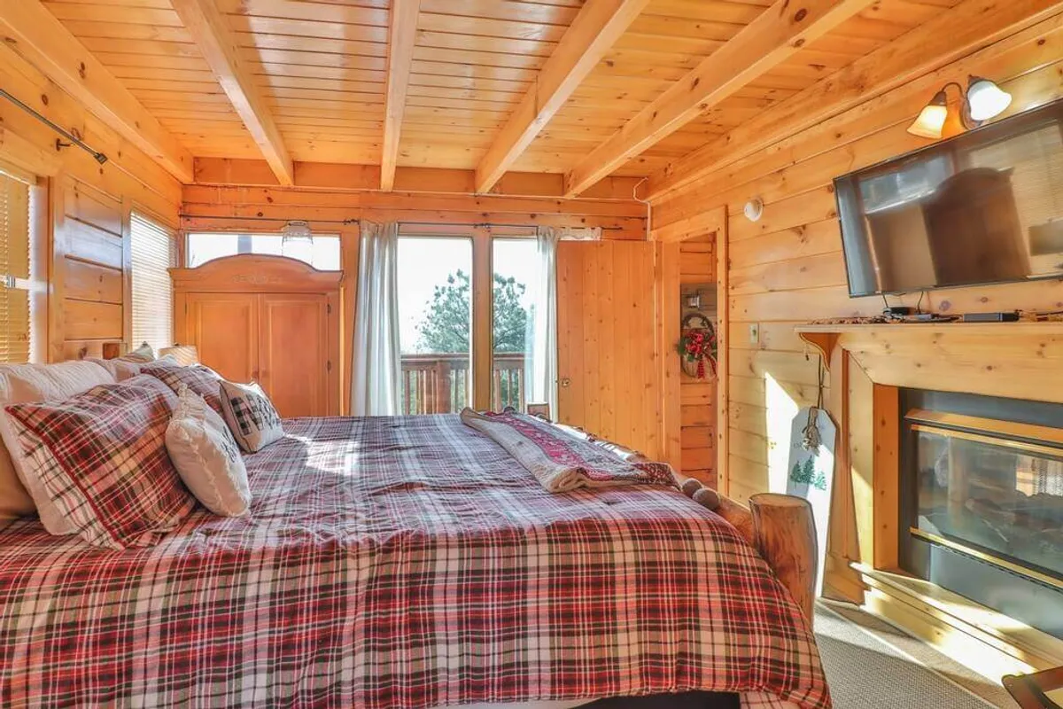 A warmly decorated bedroom with a plaid bedspread, large windows that let in abundant natural light, wooden ceilings, and a cozy fireplace. The room exudes comfort and coziness, ideal for unwinding after an adventurous day.