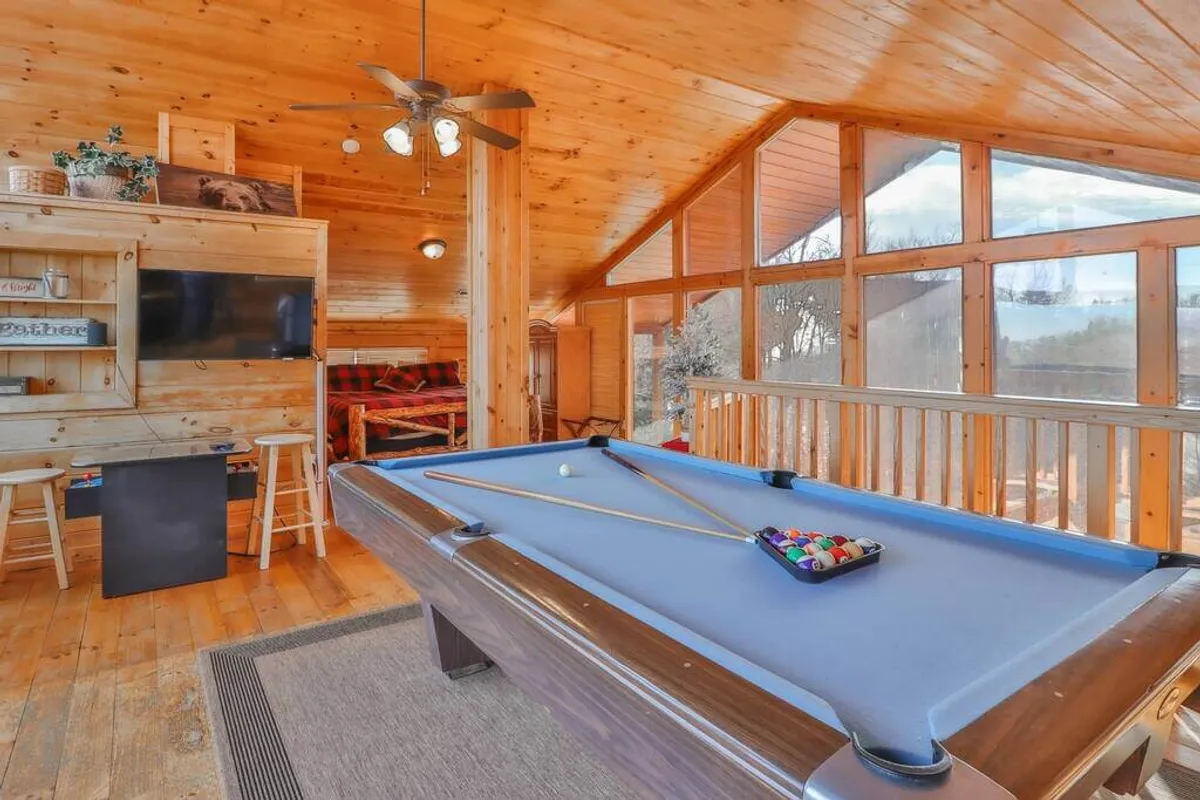 This image captures a spacious game room featuring a pool table, large windows that let in ample natural light, rich wooden walls, and a TV unit. The atmosphere is inviting and perfectly suited for socializing or engaging in friendly competition, with the pool table and TV as central elements.