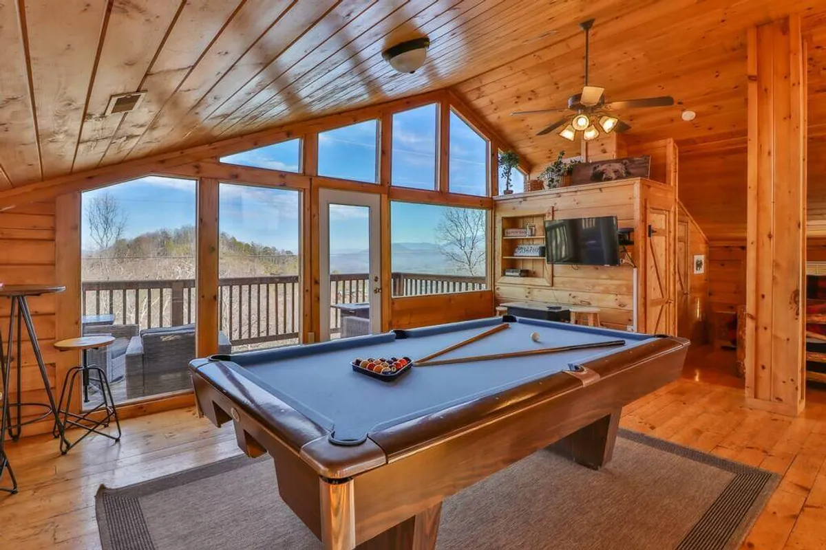 Inside the cabin, a game room features a pool table positioned before large windows that look out onto the natural surroundings. Wooden walls and a cozy sitting area enhance the friendly, relaxed vibe, creating the perfect spot for indoor entertainment and socializing with friends.