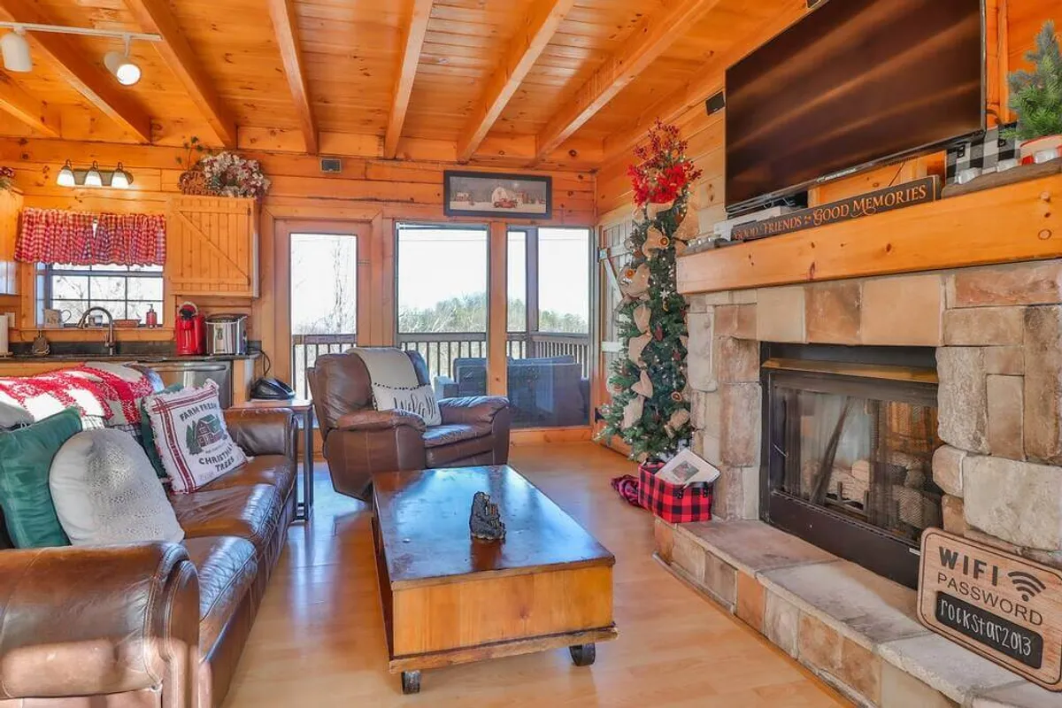 A cozy living space featuring a stone fireplace, comfortable leather sofas, a wooden coffee table, and a decorated Christmas tree. Large windows frame the area, bringing in natural light and views of the outdoors. The mood is festive yet suitable for year-round gatherings.