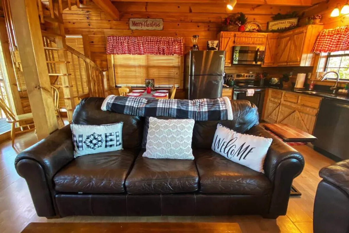 A warmly decorated living room with a brown leather couch adorned with decorative pillows, a wooden cabinet, and a dining table in the background. Large windows allow plenty of natural light in, and the space is arranged for relaxing, entertaining, and enjoying family time.